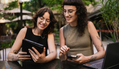 Two smiling Gen Z women at an outdoor cafe using a laptop and tablet to research home buying tips.