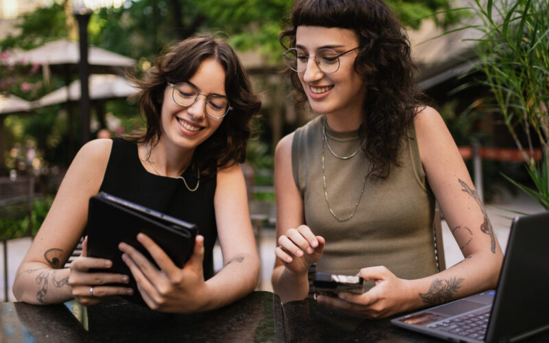 Two smiling Gen Z women at an outdoor cafe using a laptop and tablet to research home buying tips.