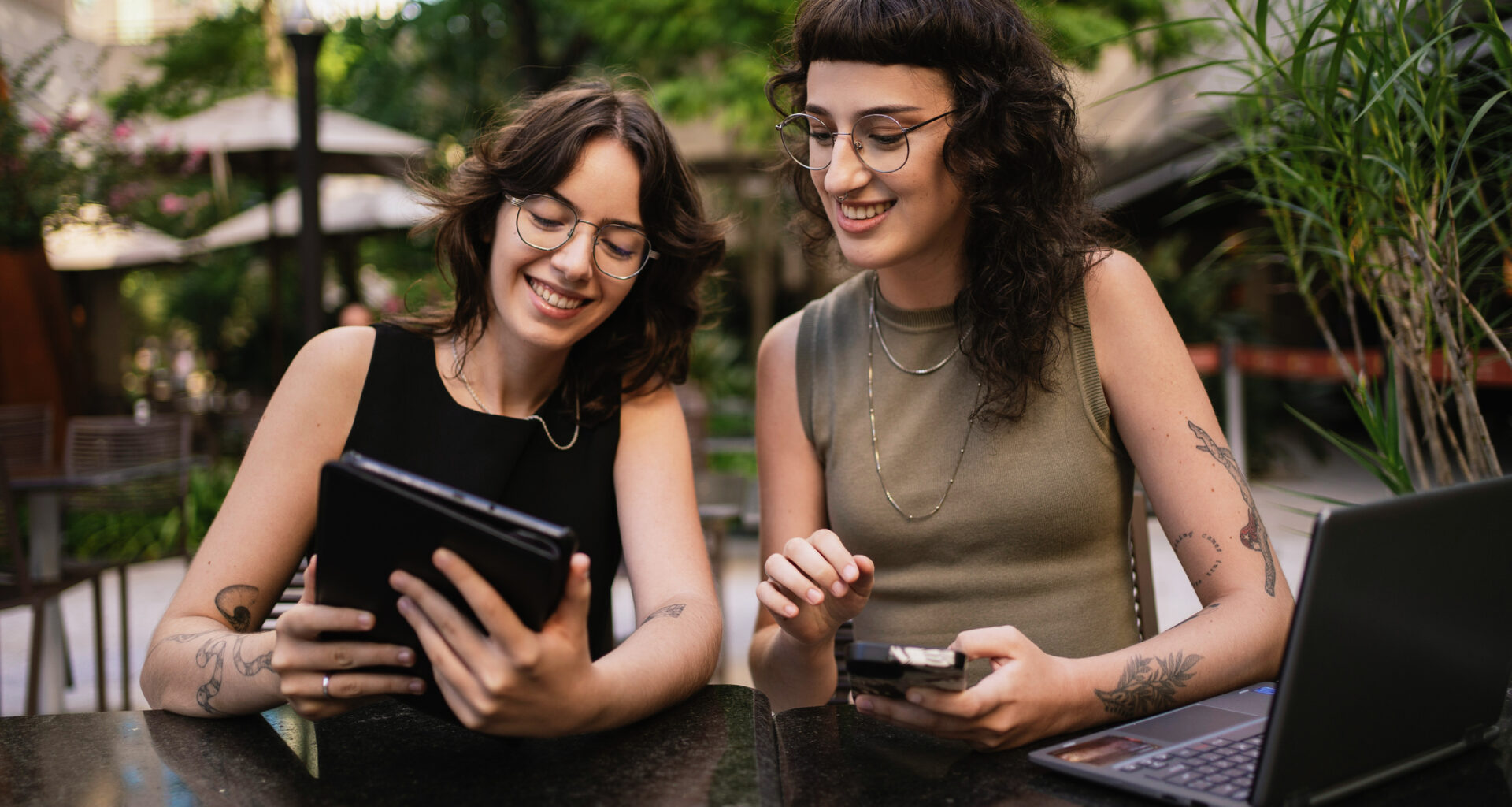 Two smiling Gen Z women at an outdoor cafe using a laptop and tablet to research home buying tips.