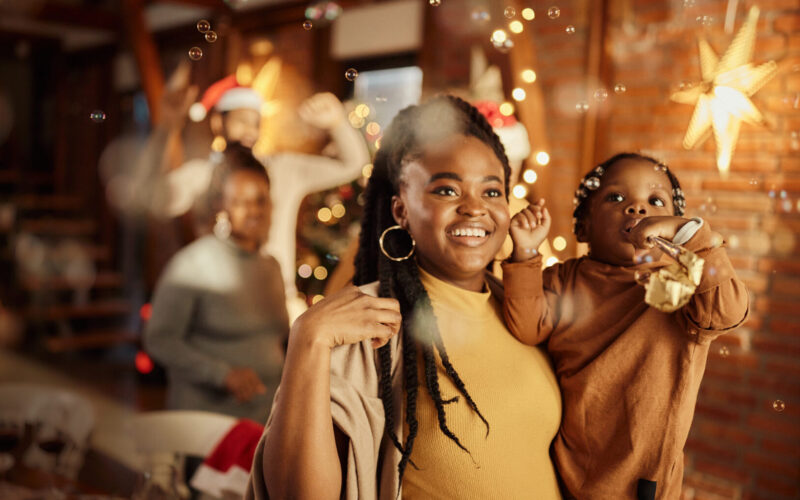 Smiling mother holding her toddler daughter celebrating New Year's Eve at home with bubbles and party horns.